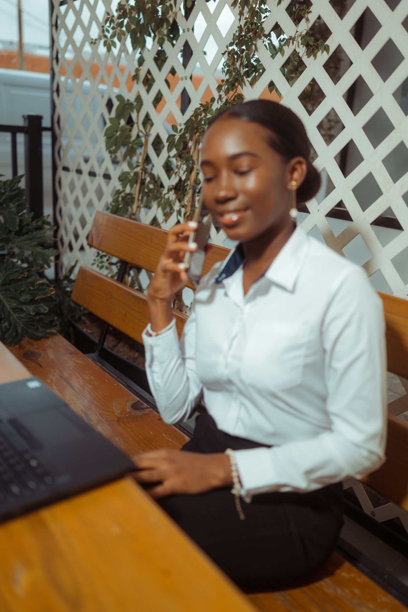 A woman sitting outdoors on a bench, holding a phone to her ear, a laptop open on the table in front of her